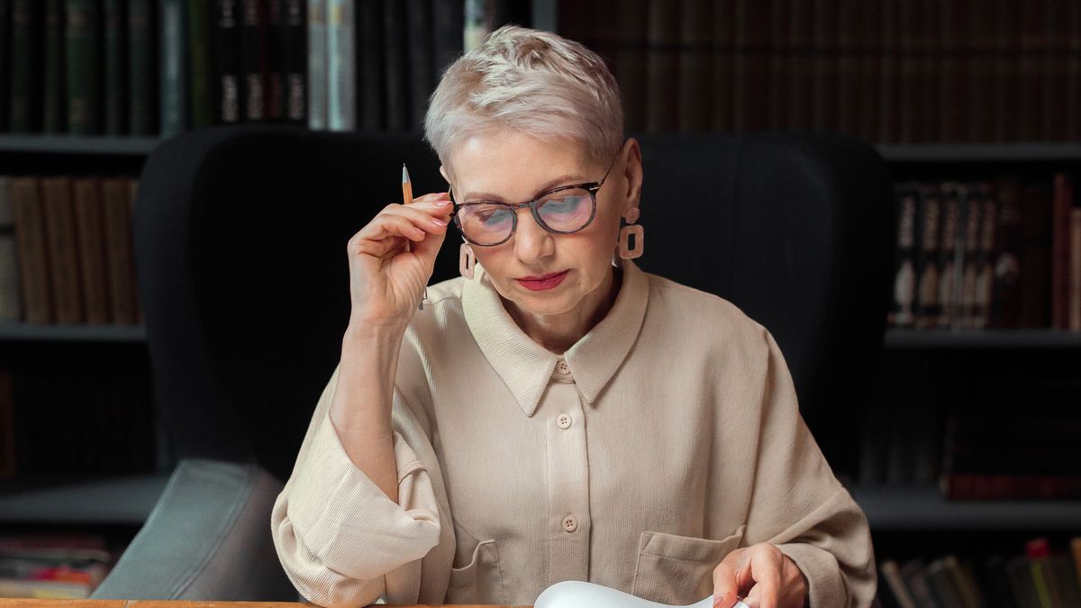 An older woman in glasses reading through paperwork at a desk, pen in hand