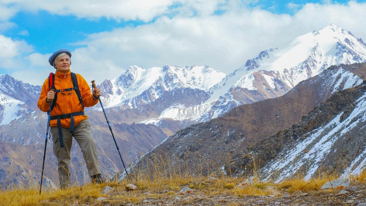 An older woman hiking alone with trekking poles below snow-capped peaks